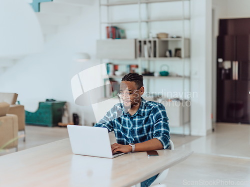 Image of African American man in glasses sitting at a table in a modern living room, using a laptop for business video chat, conversation with friends and entertainment