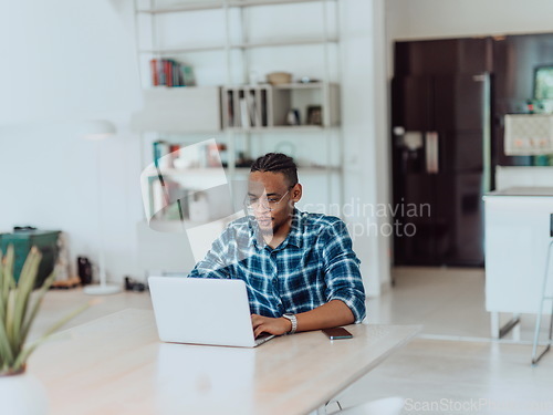 Image of African American man in glasses sitting at a table in a modern living room, using a laptop for business video chat, conversation with friends and entertainment