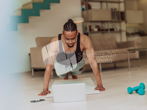 Image of Young African American man working push-ups in the living room while watching online training on laptop