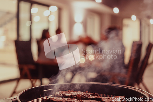 Image of Close-up photo of delicious meat being grilled. In the background, friends and family are sitting and waiting for a meal