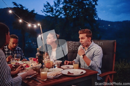 Image of A group of young diverse people having dinner on the terrace of a modern house in the evening. Fun for friends and family. Celebration of holidays, weddings with barbecue.