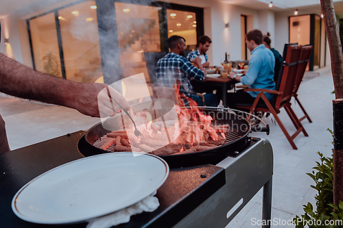 Image of A group of friends and family barbecue together in the evening on the terrace in front of a large modern house