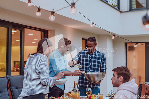 Image of A group of friends and family barbecue together in the evening on the terrace in front of a large modern house