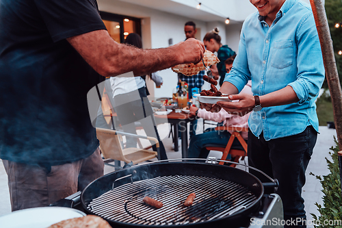 Image of A group of friends and family barbecue together in the evening on the terrace in front of a large modern house