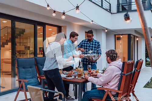 Image of A group of young diverse people having dinner on the terrace of a modern house in the evening. Fun for friends and family. Celebration of holidays, weddings with barbecue.