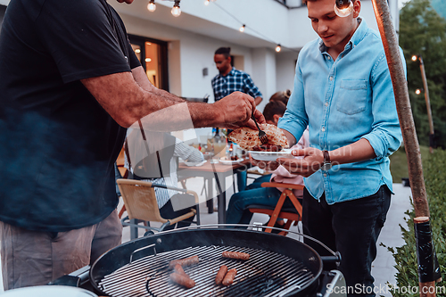 Image of A group of friends and family barbecue together in the evening on the terrace in front of a large modern house