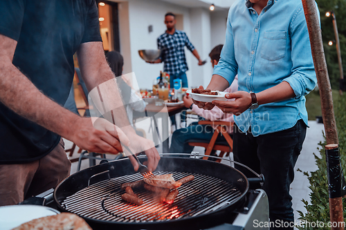 Image of A group of friends and family barbecue together in the evening on the terrace in front of a large modern house