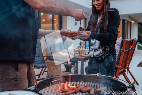 Image of A group of friends and family barbecue together in the evening on the terrace in front of a large modern house