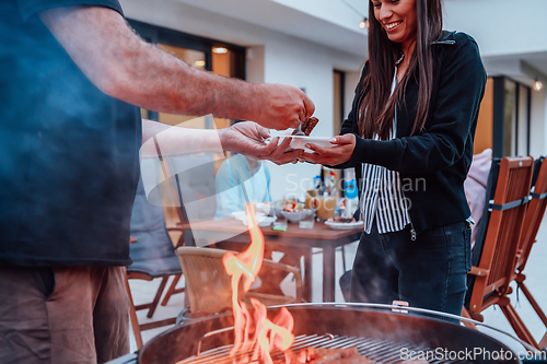 Image of A group of friends and family barbecue together in the evening on the terrace in front of a large modern house