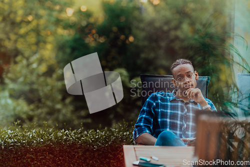 Image of An African American man sitting in front of the house on a modern terrace and stares thoughtfully at the camera