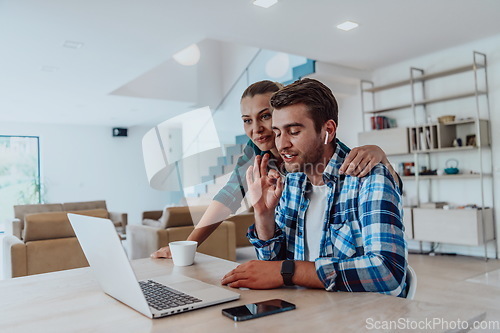 Image of A young married couple is talking to parents, family and friends on a video call via a laptop while sitting in the living room of their modern house