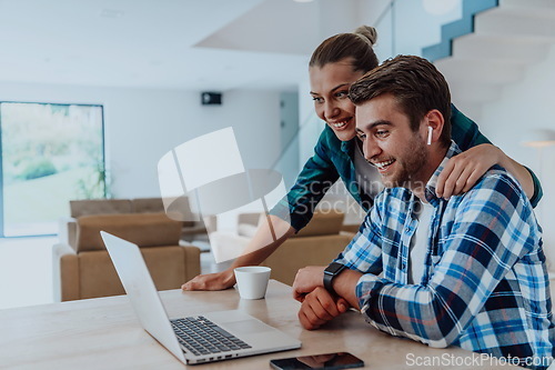 Image of A young married couple is talking to parents, family and friends on a video call via a laptop while sitting in the living room of their modern house