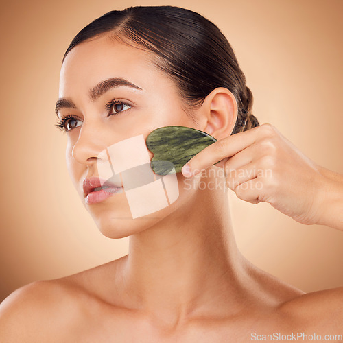 Image of Beauty, gua sha and face of woman for skincare, facial treatment and wellness with spa stone. Salon, dermatology and girl on brown background with cosmetics, face massage and luxury tools in studio