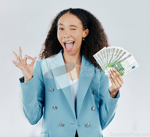 Image of Portrait, wink and cash with a business woman in studio on a gray background making a perfect hand gesture. Funny face, finance and money with a female employee holding euro bills for investment