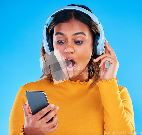 Image of Headphones, cellphone and shocked woman in a studio listening to a podcast, music or radio. Surprise, technology and female model streaming an album, song or playlist with a phone by blue background.