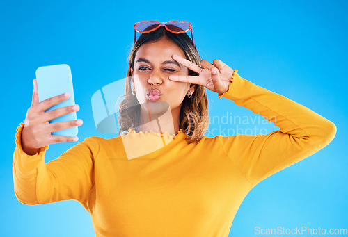 Image of Selfie, studio and peace sign of a woman influencer taking a profile picture for social media. Isolated, blue background and silly hands gesture of a gen z female with a kiss feeling happy and fun