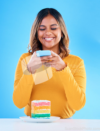 Image of Woman, cake and studio with a phone for social media picture with a smile and excited to eat. Happy female person on blue background with rainbow color dessert for birthday or influencer celebration