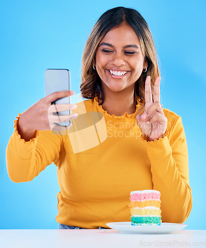 Image of Woman, cake and peace selfie in studio for social media with a smile and excited to eat. Happy, female person and hand emoji on blue background with rainbow color dessert for birthday celebration