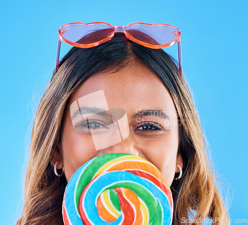 Image of Portrait, giant lollipop and a woman on a blue background in studio wearing heart glasses for fashion. Face, candy and sweet with an attractive young female eating a snack while feeling happy