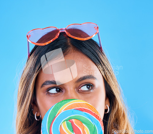 Image of Thinking, lollipop and a woman on a blue background in studio wearing heart glasses for fashion. Idea, candy and sweet with an attractive young female eating a giant snack while feeling thoughtful