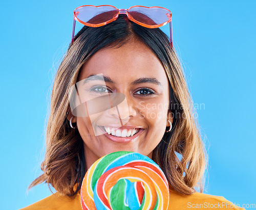 Image of Portrait, lollipop and a woman smiling on a blue background in studio wearing heart glasses for fashion. Face, candy and sweet with a happy young female eating a giant snack while feeling positive