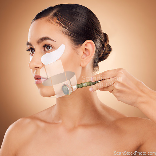 Image of Woman beauty, eye patch and face roller for studio dermatology, healthcare or skincare treatment. Facial massage product, dermatology or female model with hyaluronic acid mask on brown background