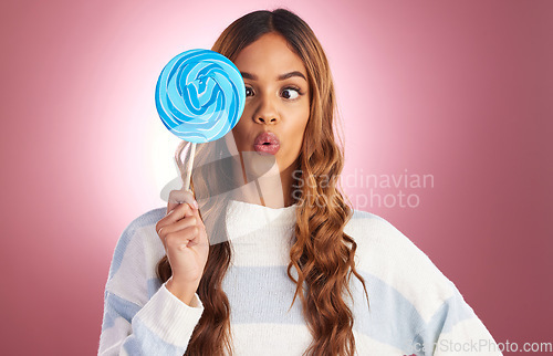 Image of Portrait, funny face and lollipop with a woman on a pink background in studio holding giant candy. Comic, comedy and sweet with a playful young female feeling silly while eating a blue sugar treat