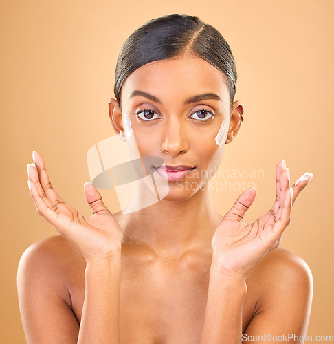 Image of Face portrait, skincare and woman with cream in studio isolated on a brown background. Dermatology, cosmetics and serious Indian female model apply lotion, creme and moisturizer for healthy skin.