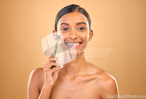 Image of Face portrait, skincare roller and woman in studio isolated on a brown background. Dermatology, facial massage and happy Indian female model with jade crystal for healthy skin treatment and beauty.