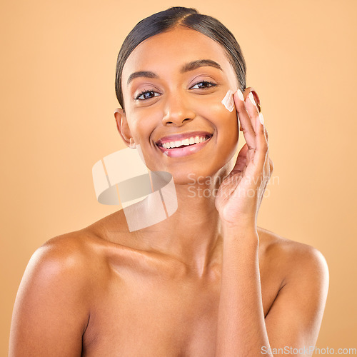 Image of Face portrait, skincare and woman with cream in studio isolated on a brown background. Dermatology smile, cosmetics and happy Indian female model apply lotion, creme and moisturizer for healthy skin.