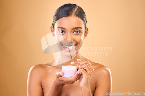 Image of Face, skincare and woman with cream jar in studio isolated on a brown background. Dermatology cosmetics, portrait and happy Indian female apply lotion, creme and moisturizer product for healthy skin.