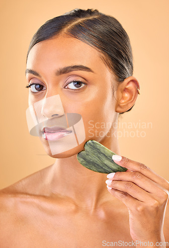 Image of Face portrait, skincare and woman with gua sha in studio isolated on a brown background. Dermatology, massage and serious Indian female model with jade crystal or stone for healthy skin treatment.