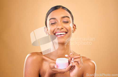 Image of Face, skincare and woman with cream jar in studio isolated on a brown background. Dermatology cosmetics, funny and happy Indian female apply lotion, creme and moisturizer product for healthy skin.