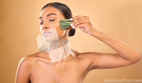 Image of Face, skincare stone and woman with eyes closed in studio isolated on a brown background. Dermatology, facial massage and Indian female model with jade crystal or gua sha for healthy skin treatment.