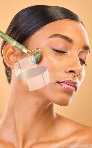 Image of Face, skincare roller and woman with eyes closed in studio isolated on a brown background. Dermatology, facial massage and Indian female model with jade crystal for healthy skin treatment and beauty.