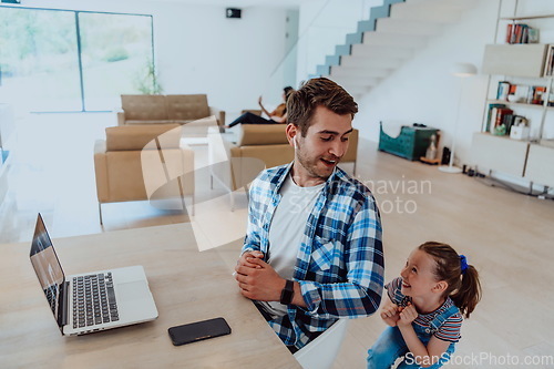 Image of Work from home. Daughter interrupting her father while he is having a business online conversation on his laptop while sitting in modern living room