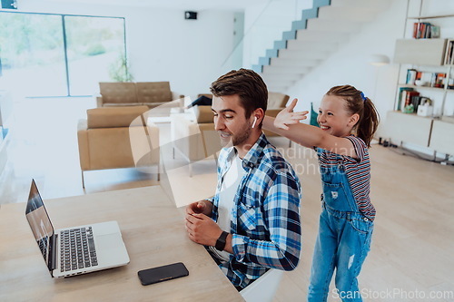 Image of Work from home. Daughter interrupting her father while he is having a business online conversation on his laptop while sitting in modern living room