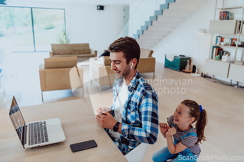 Image of Work from home. Daughter interrupting her father while he is having a business online conversation on his laptop while sitting in modern living room