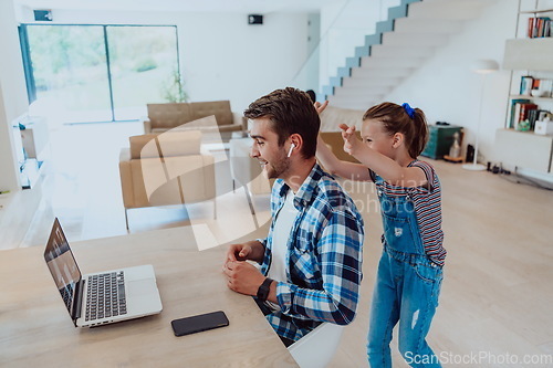 Image of Work from home. Daughter interrupting her father while he is having a business online conversation on his laptop while sitting in modern living room