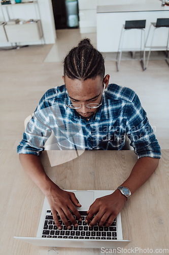 Image of African American man in glasses sitting at a table in a modern living room, using a laptop for business video chat, conversation with friends and entertainment