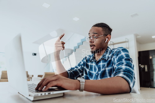 Image of African American man in glasses sitting at a table in a modern living room, using a laptop for business video chat, conversation with friends and entertainment