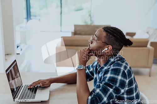 Image of African American man in glasses sitting at a table in a modern living room, using a laptop for business video chat, conversation with friends and entertainment