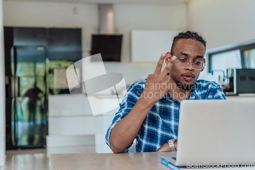Image of African American man in glasses sitting at a table in a modern living room, using a laptop for business video chat, conversation with friends and entertainment