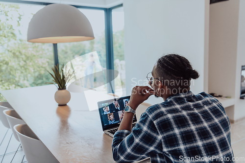 Image of African American man in glasses sitting at a table in a modern living room, using a laptop and smartphone for business video chat, conversation with friends and entertainment