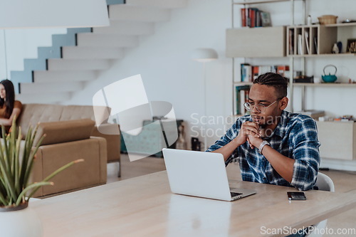 Image of African American man in glasses sitting at a table in a modern living room, using a laptop for business video chat, conversation with friends and entertainment