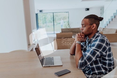 Image of African American man in glasses sitting at a table in a modern living room, using a laptop for business video chat, conversation with friends and entertainment