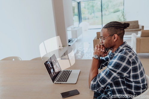 Image of African American man in glasses sitting at a table in a modern living room, using a laptop for business video chat, conversation with friends and entertainment