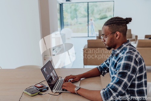 Image of African American man in glasses sitting at a table in a modern living room, using a laptop for business video chat, conversation with friends and entertainment