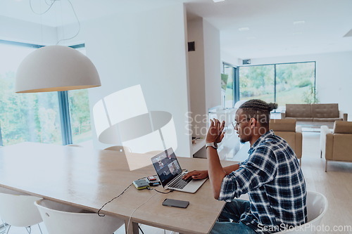 Image of African American man in glasses sitting at a table in a modern living room, using a laptop for business video chat, conversation with friends and entertainment