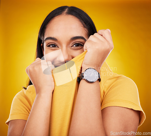 Image of Portrait, shy and funny with a woman on a yellow background in studio pulling on her t-shirt. Face, fashion or playful and an attractive young female covering her mouth with trendy clothes for style